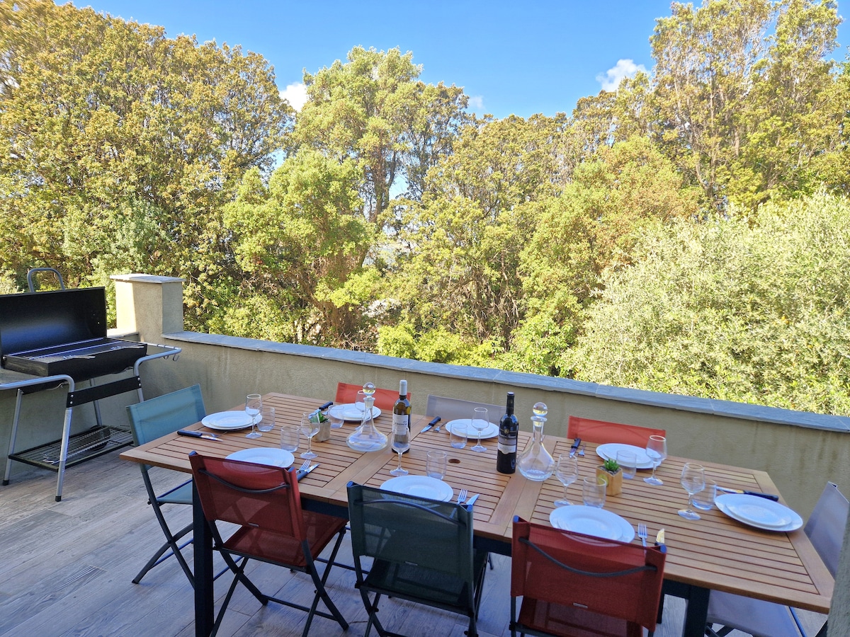 An outdoor dining space is presented with a wooden table set for six, showcasing dishes and glassware. Colorful chairs surround the table. A barbecue grill is positioned nearby, with a backdrop of lush greenery and trees, creating a serene environment for meals.