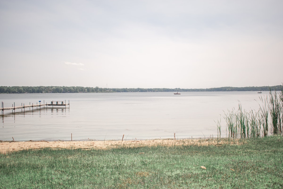 A serene view of Bass Lake presents calm waters meeting a sandy shoreline, bordered by green grasses. In the distance, a dock extends into the lake, with small boats visible. Lush greenery outlines the far shore, creating a tranquil natural setting.