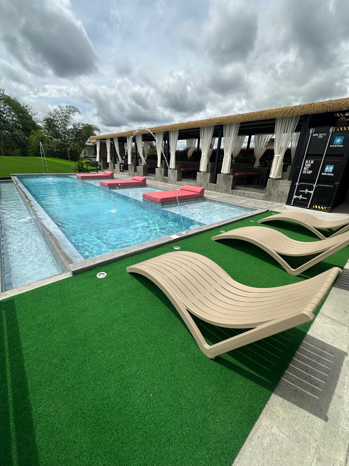 An inviting pool area features a shimmering blue pool surrounded by green grass. Multiple lounge chairs are positioned for relaxation, while water features add a dynamic element. Covered seating areas with draped curtains provide shaded options for enjoying the outdoor space.