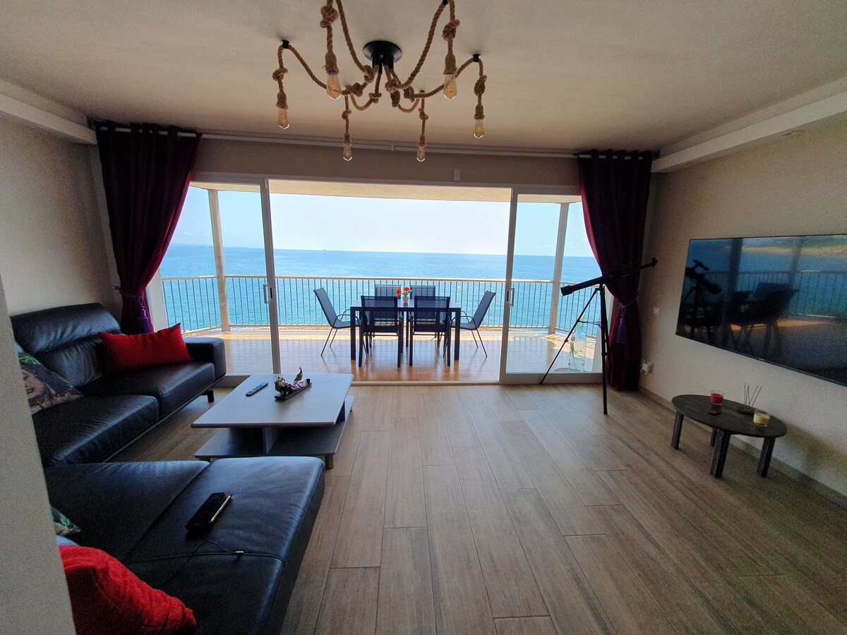 A spacious living room features a large glass sliding door that opens to stunning sea views. Dark curtains frame the window. A comfortable sectional sofa with decorative pillows is positioned alongside a modern coffee table. A dining table for six is visible on the terrace.