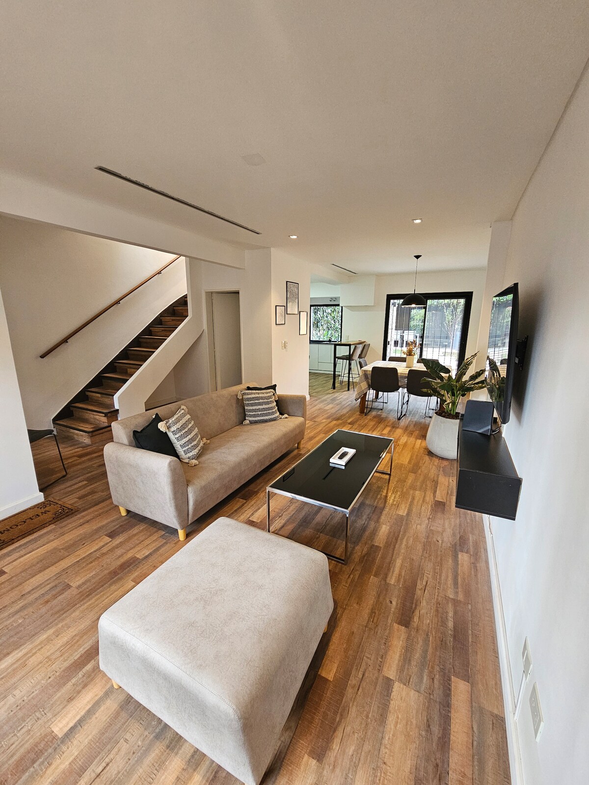 An open living and dining area is presented, featuring modern furnishings including a light-colored sofa, a glass coffee table, and a plush ottoman. Natural light fills the space, highlighting the wooden flooring. A staircase is visible in the background, leading to the upper level.