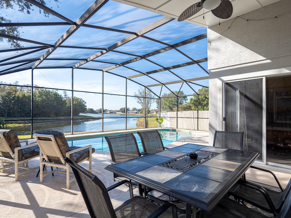 An enclosed patio space features a dining table surrounded by chairs, with views of a tranquil waterway. Comfortable seating areas are situated nearby, highlighted by the natural light filtering through the screened structure. The pool is visible through the enclosure, enhancing the outdoor living experience.