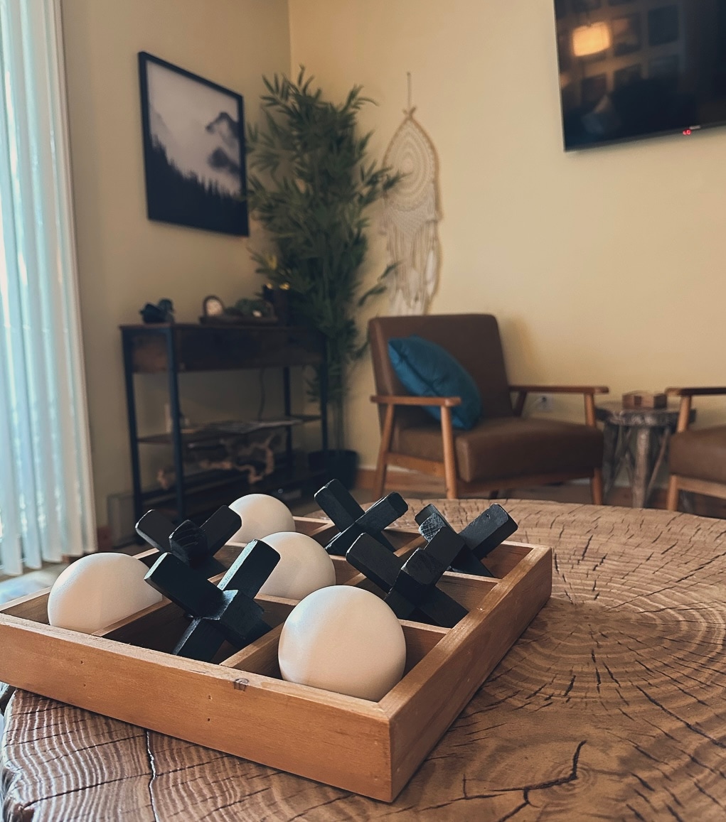 A wooden game board is placed on a rustic round table, featuring a tic-tac-toe design with white and black pieces. The background includes a cozy seating area with two brown chairs, a small console table, and decorative wall art, adding a serene touch to the space.