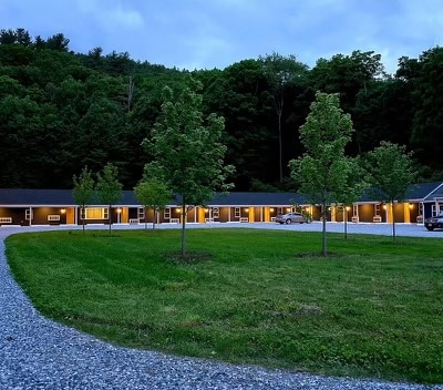 A serene landscape showcases a mid-century motel surrounded by lush trees under twilight. A circular gravel driveway leads to the entrance, where soft lighting highlights the rooms. Well-kept grass adds a touch of nature to the tranquil setting.