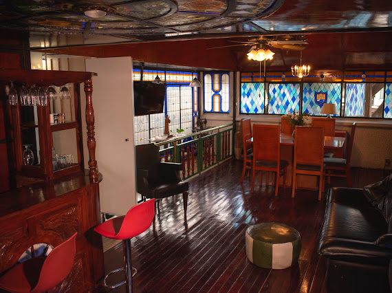 A stylish living area features stained glass windows that allow natural light to illuminate the space. A wooden bar with glassware is positioned nearby, while a dining table with six chairs adds to the cozy atmosphere. Dark wooden floors enhance the warm ambiance.