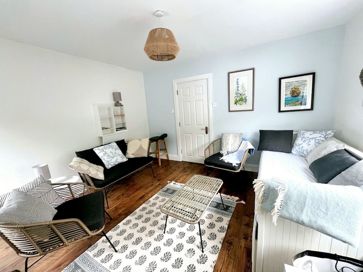 A well-furnished living area features a cozy layout with a sofa, two armchairs, and a daybed. Soft textiles and a patterned rug add comfort. Two framed artworks adorn the walls, while natural light brightens the room through a nearby window, creating a calm ambiance.