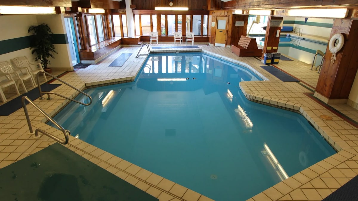 An indoor pool area features a spacious layout with clear blue water. Natural light floods through large windows, highlighting the tiled surroundings. Seating is available along one side, enhancing the inviting atmosphere for relaxation and leisure activities.