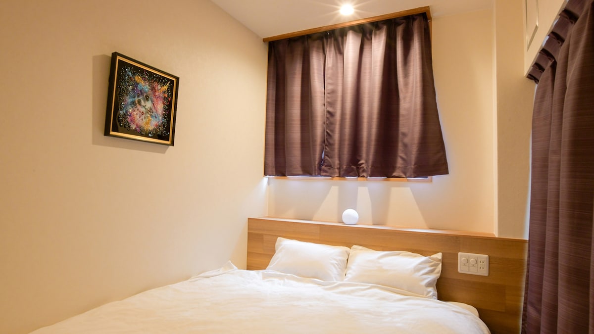 A cozy bedroom features a spacious bed with white linens positioned against a light wooden headboard. A piece of artwork is displayed above the bed, and soft lighting emanates from wall fixtures. Dark curtains frame the window, allowing for privacy and comfort.