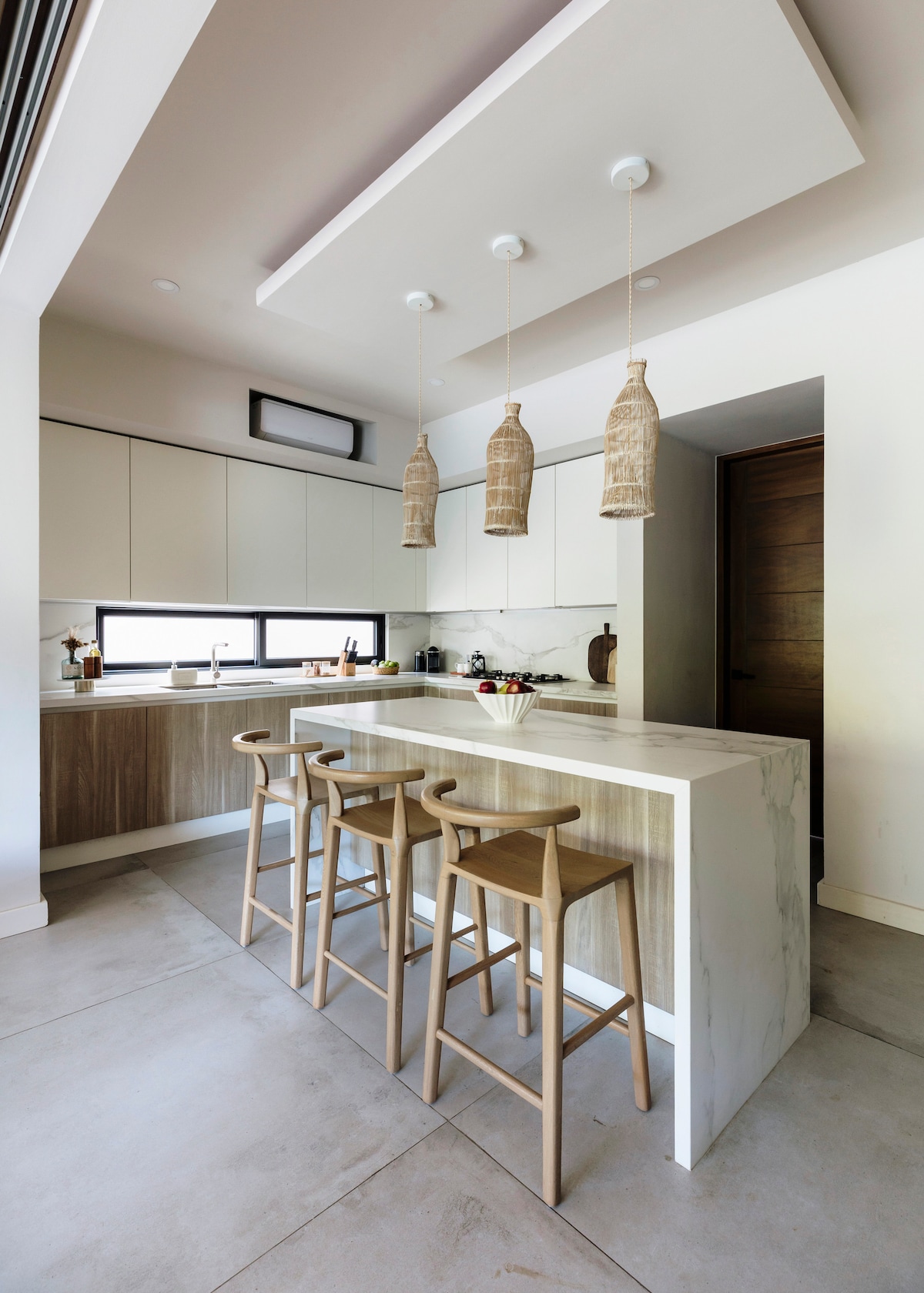 The kitchen features modern cabinetry and a light-colored marble island with three wooden bar stools. Pendant lights hang above the island, providing subtle illumination. Large windows allow natural light to fill the space, highlighting the sleek design and functionality of the kitchen.