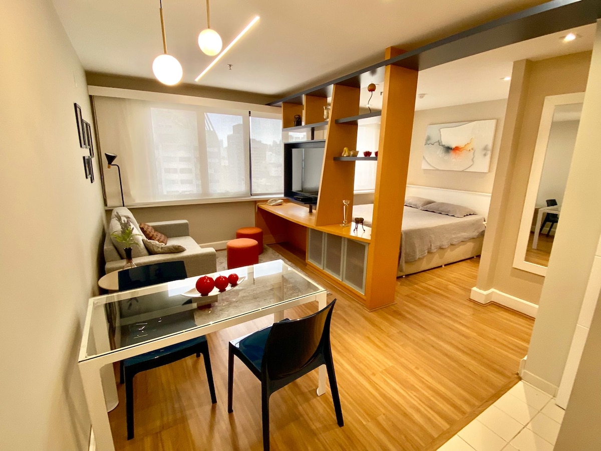 A contemporary living space features a cozy seating area with a sofa and an orange accent table. A dining table is set nearby, accompanied by black chairs. In the background, a spacious bed is positioned next to a flat-screen TV and light wood cabinetry, creating a functional layout.