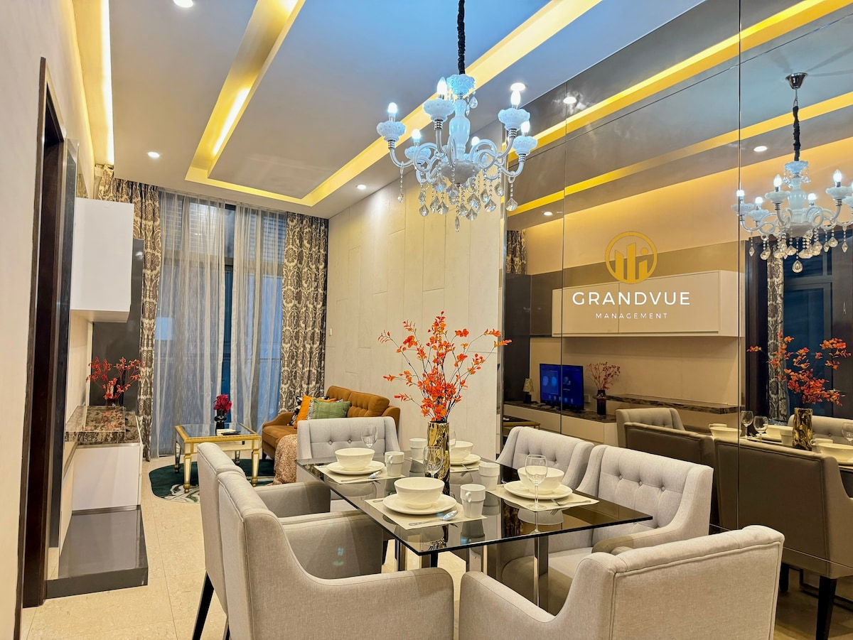 A dining area is visible with a glass table set for dining, surrounded by four upholstered chairs. A cozy seating area features a plush sofa adorned with pillows. Tall glass windows allow natural light to fill the space, while modern lighting fixtures hang from the ceiling.