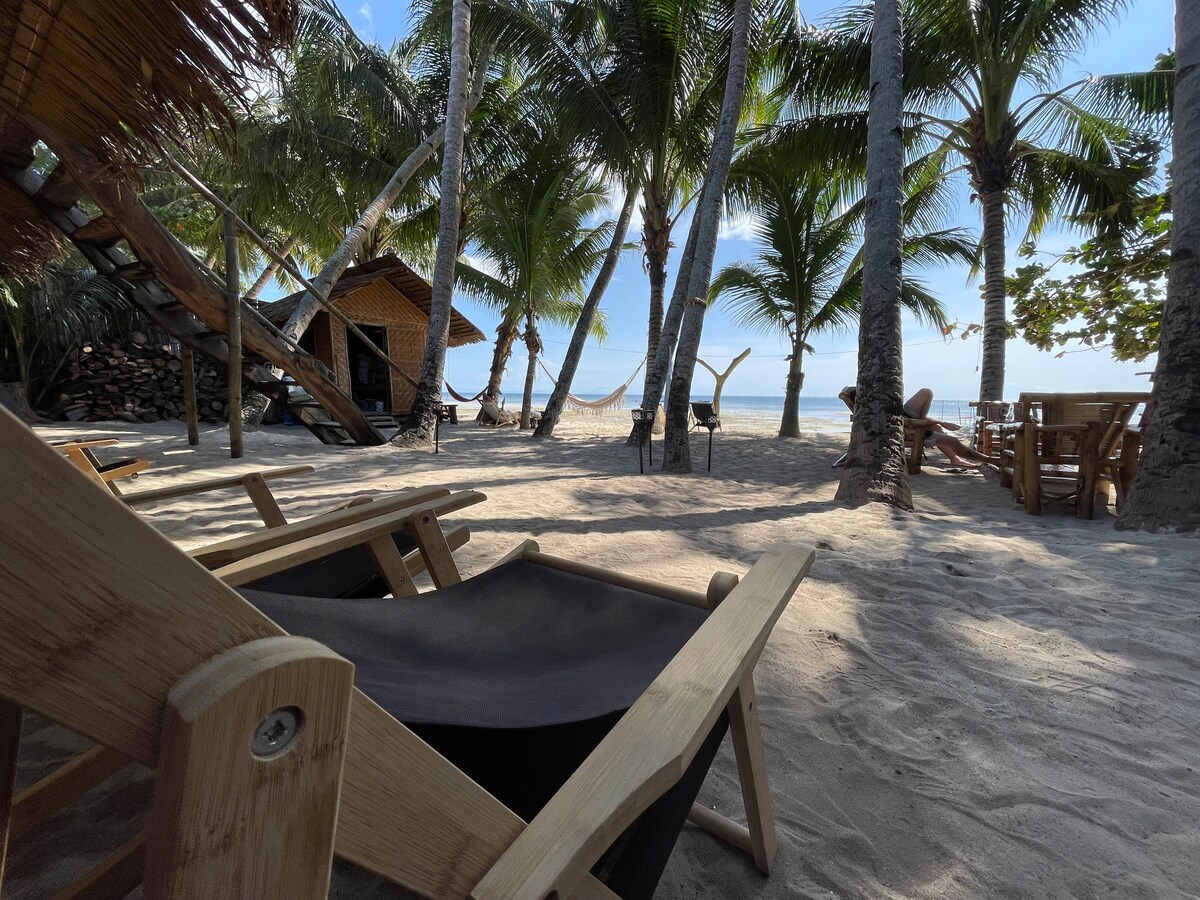A sandy beach area is lined with tall palm trees, providing shade and a tropical feel. Several wooden lounge chairs are positioned for relaxation. In the background, a hammock is visible, along with a small wooden structure, and a dining table can be seen to the side.