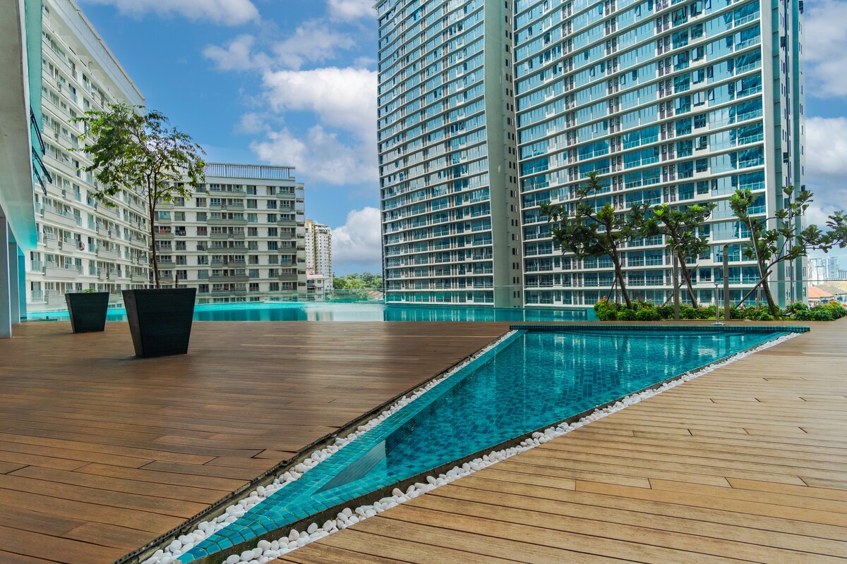 An outdoor swimming pool is framed by wooden decking and modern high-rise buildings. The pool features a unique design, with a narrow channel lined with small white stones, creating a tranquil atmosphere amidst the urban landscape. Lush greenery is present, enhancing the setting.