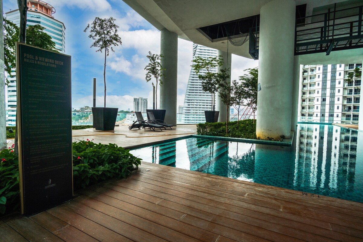 An inviting infinity pool is framed by wood decking and lush greenery. Sun loungers are positioned nearby, offering a space for relaxation. The backdrop features modern high-rise buildings and clear blue skies, creating a serene environment for guests to enjoy.