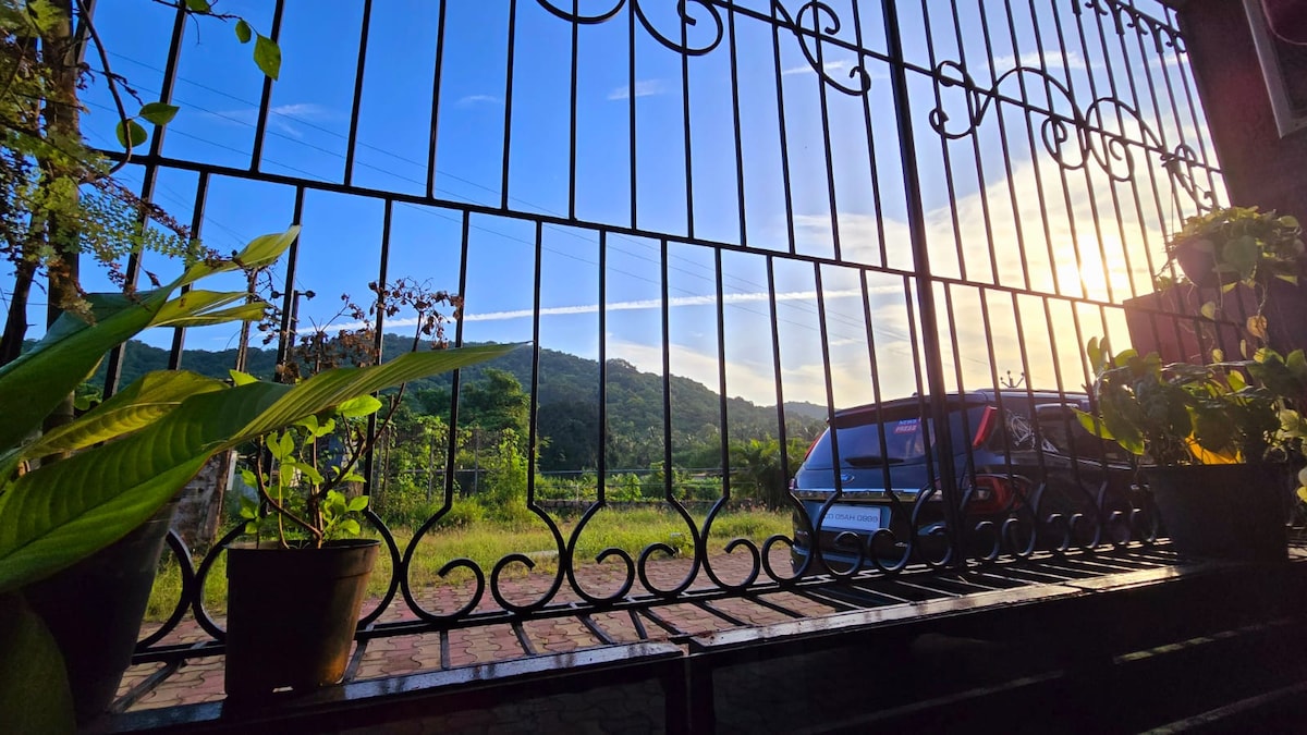 A view from the balcony showcases lush green hills under a clear blue sky, framed by a decorative iron railing. Potted plants populate the balcony space, enhancing the natural setting. A vehicle is parked just outside, complementing the serene surroundings.