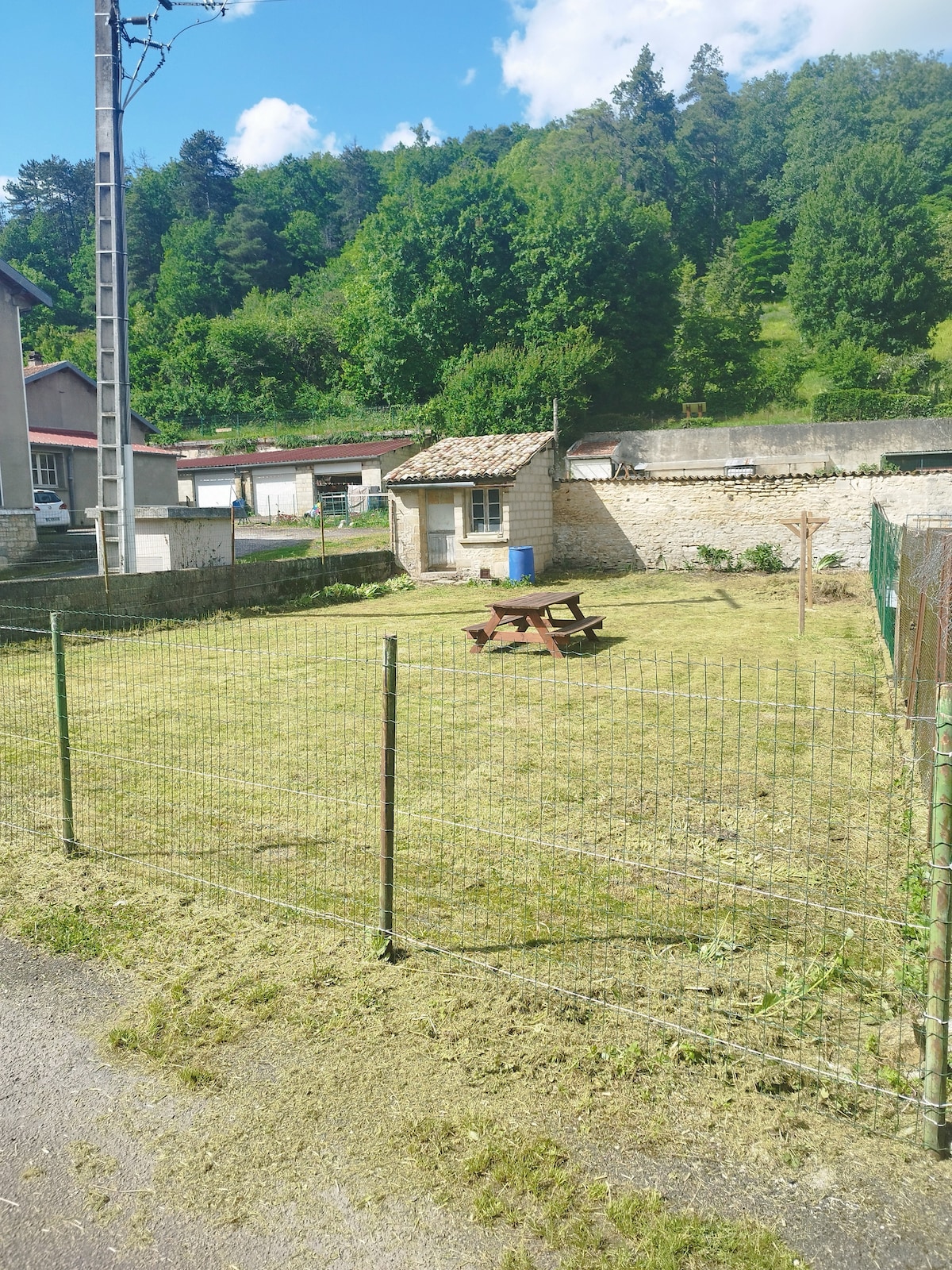 A fenced outdoor area features a grassy lawn with a picnic table at its center. A small, rustic stone building is visible in the background, surrounded by trees and greenery. The setting is bright, illuminated by natural sunlight.