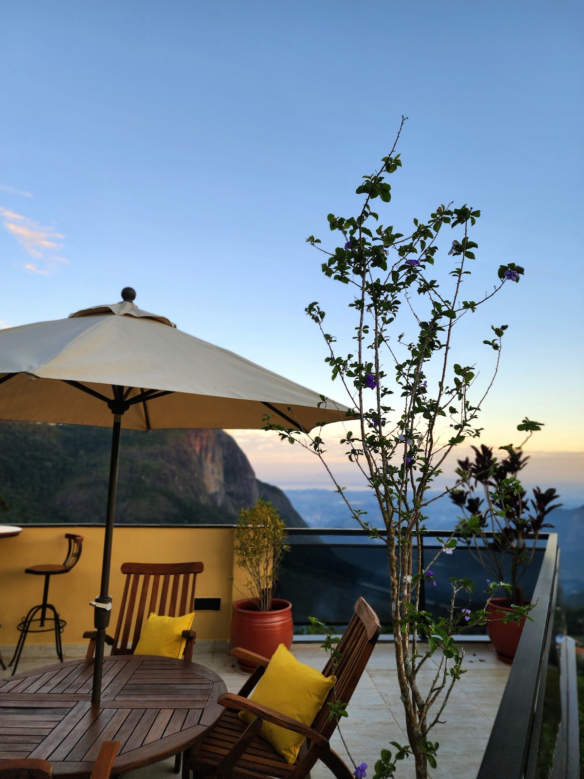 A private terrace features wooden dining furniture and a large umbrella providing shade. Potted plants and greenery adorn the area, complementing the scenic mountain views in the background. The sky is clear, enhancing the serene outdoor space.