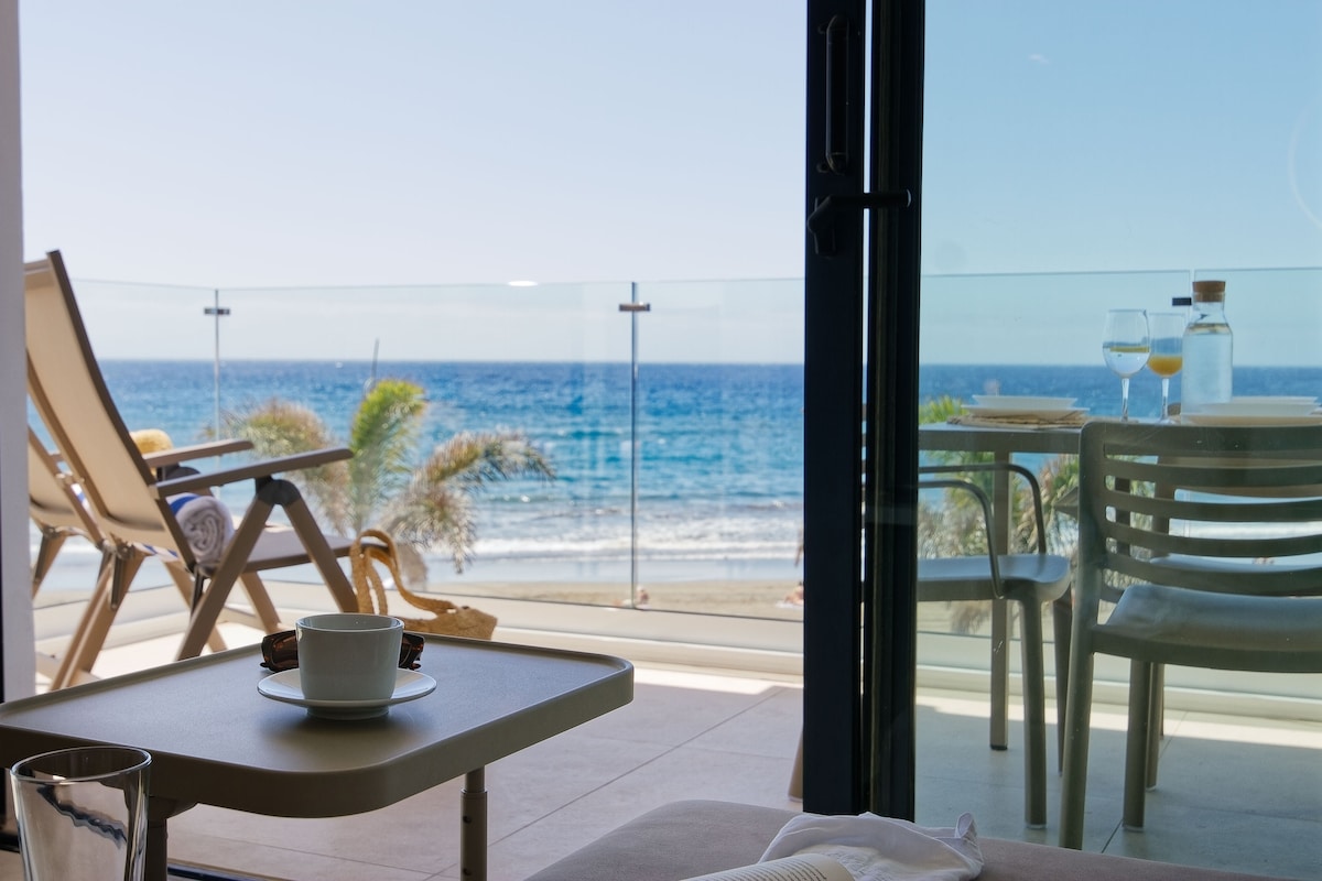 A seaside view is captured through large glass doors, revealing a terrace with comfortable chairs overlooking the ocean. A small table displays a cup and a glass, while gentle waves can be seen in the distance, providing a sense of tranquility.