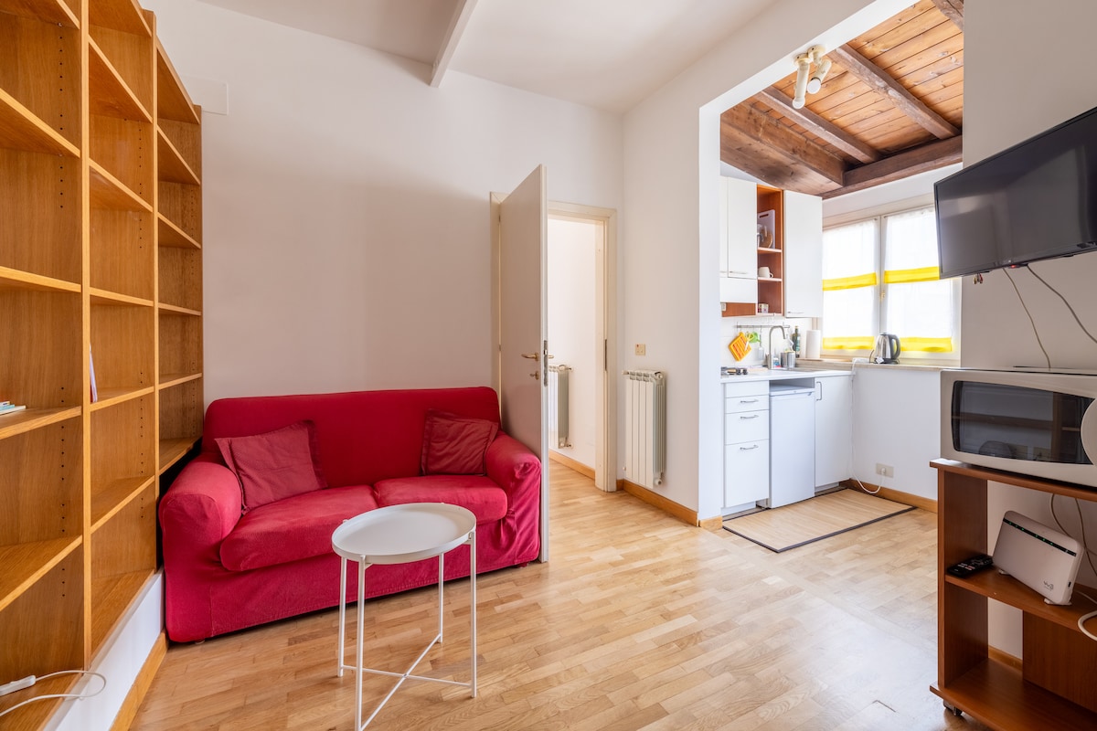 The living area features a soft pink sofa, accompanied by a small white table. A wooden bookshelf lines one wall, while a kitchenette is visible in the background, showcasing a stove and sink. Natural light enters through windows with yellow accents, illuminating the space.