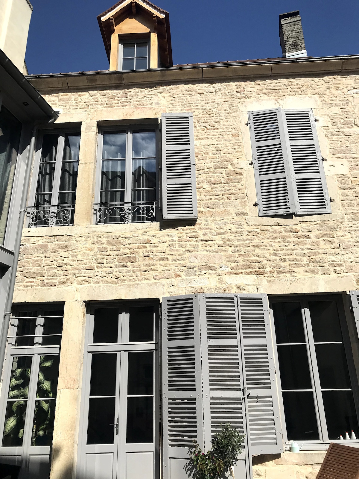 The image showcases a charming stone exterior with multiple windows, including shutters painted in gray. A sloped roof with wooden accents adds character, while a decorative balcony can be seen above. Light streams in, highlighting the texture of the stone facade.