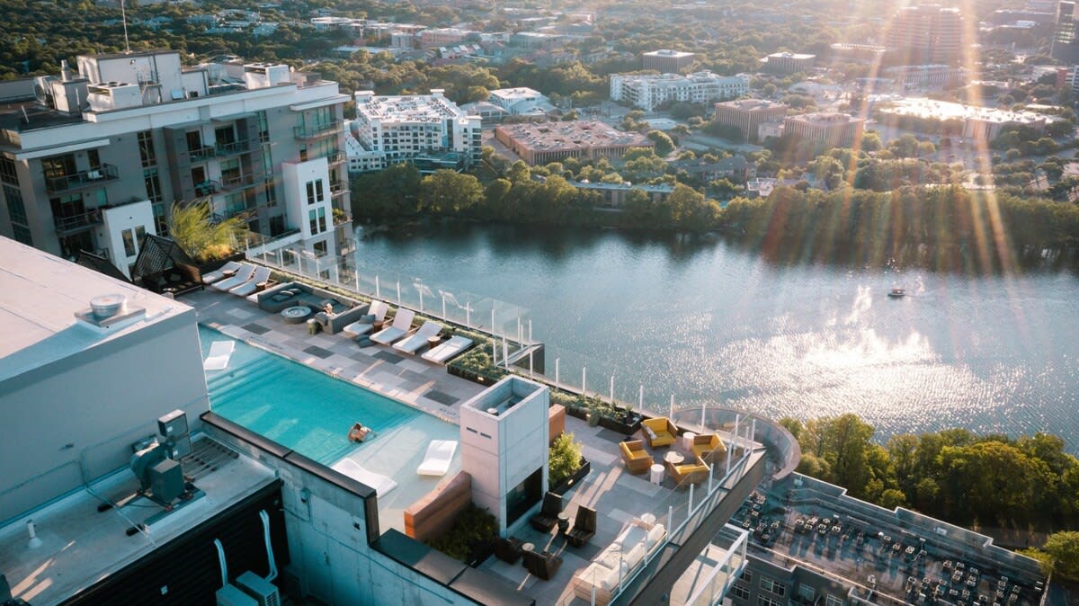 The rooftop pool offers a panoramic view of the surrounding landscape and Lady Bird Lake. Lounge chairs are arranged around the pool area, complemented by stylish seating options. The vibrant city skyline is visible in the background, with sun rays reflecting off the water's surface.