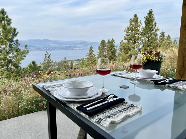 A glass table set for two features plates, cutlery, and glasses of red wine. The table overlooks a vibrant wildflower garden, with scenic views of Lake Okanagan and the surrounding mountains in the background.
