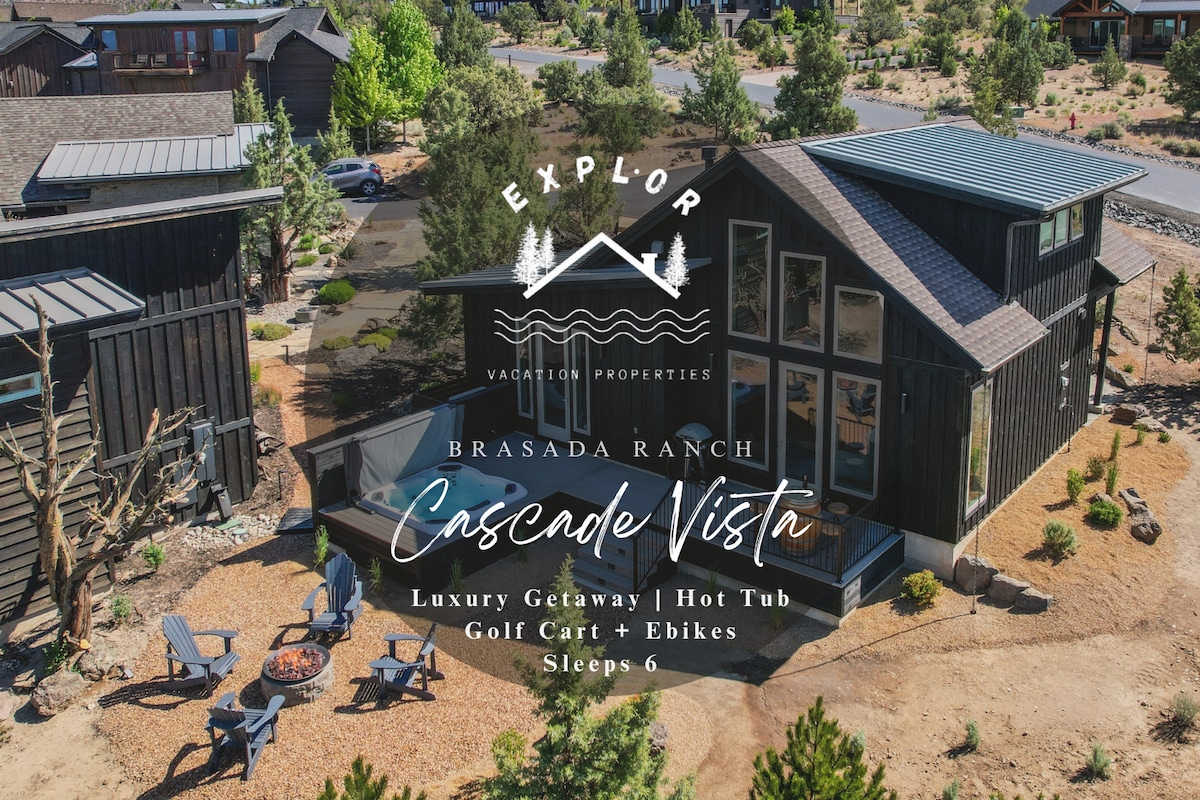 A modern casita features large glass windows, reflecting natural light and surrounding greenery. Outdoor seating with several chairs encircles a fire pit, with rustic pathways leading through the landscape. The structure presents a blend of dark wood accents and contemporary design elements amidst a serene setting.