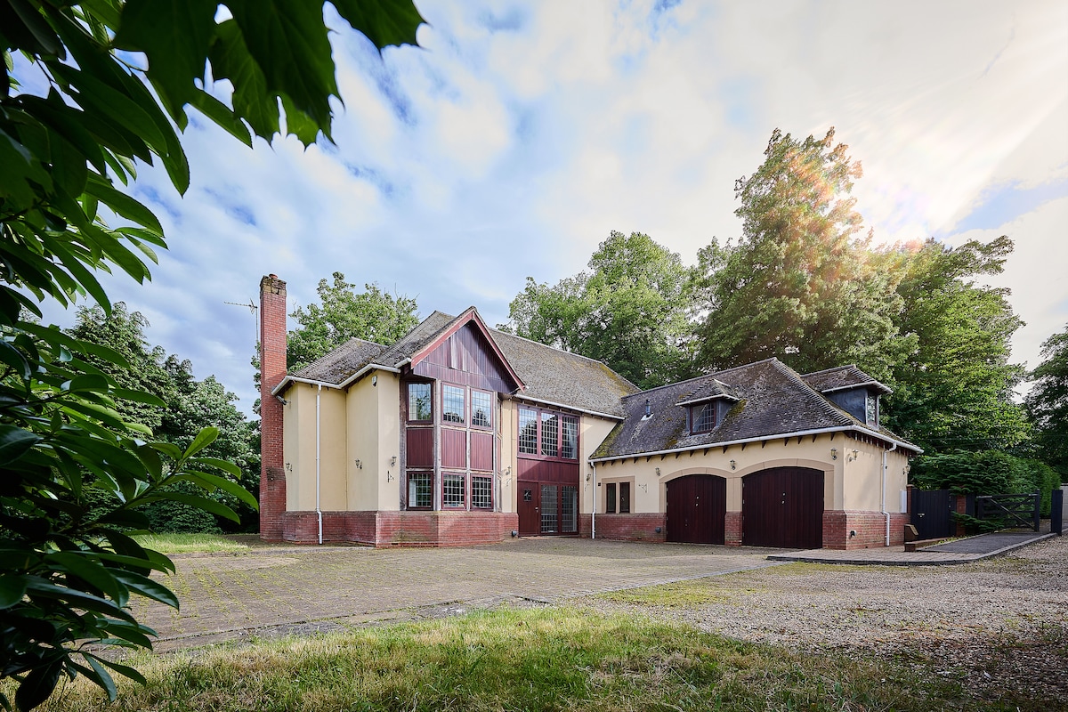 The exterior of the spacious property is captured, showcasing a two-story home with a combination of red and cream siding. Large windows are featured, allowing natural light to illuminate the interior. A gravel driveway leads to the double garage, surrounded by greenery.