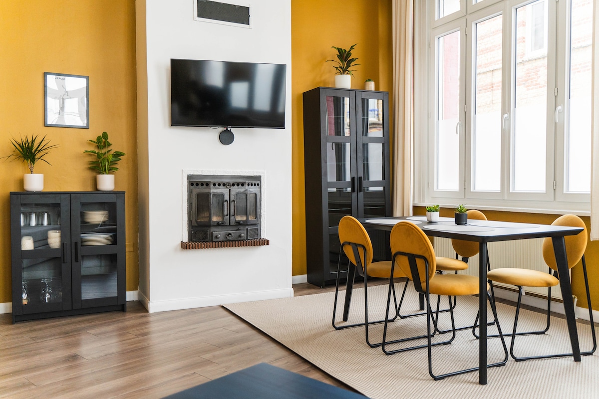 The living area features a warm yellow wall with a flat-screen television mounted above a modern fireplace. A dining table with yellow chairs accommodates four. Glass-fronted cabinets display decorative items and plants, while natural light fills the space through multiple large windows.