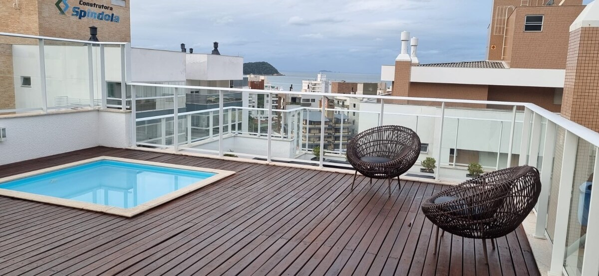A rooftop terrace presents a small pool surrounded by wooden decking. Two brown woven chairs offer seating with a view of the distant beach and coastal buildings. The clear glass railing provides an unobstructed perspective of the outdoor space.