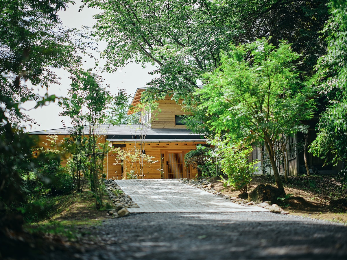 A serene approach leads to a two-story wooden building, surrounded by lush greenery. The pathway is lined with smooth stones, guiding the way to the entrance. Natural light filters through the canopy of trees, creating a peaceful ambiance.