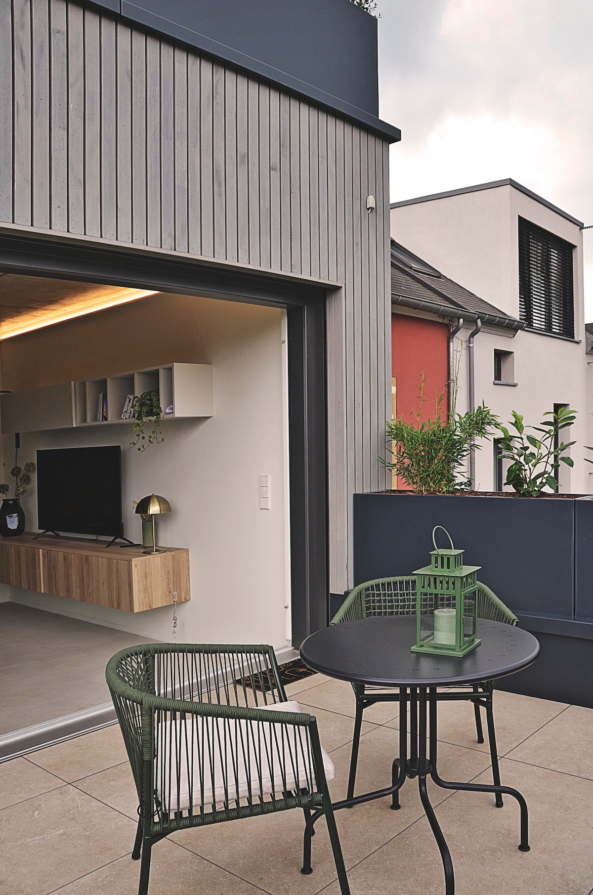 An outdoor balcony space is visible, featuring a round black table accompanied by two green chairs. A lantern rests on the table, while the sleek wooden exterior of the building is contrasted by clean, modern lines and subtle greenery in the background.
