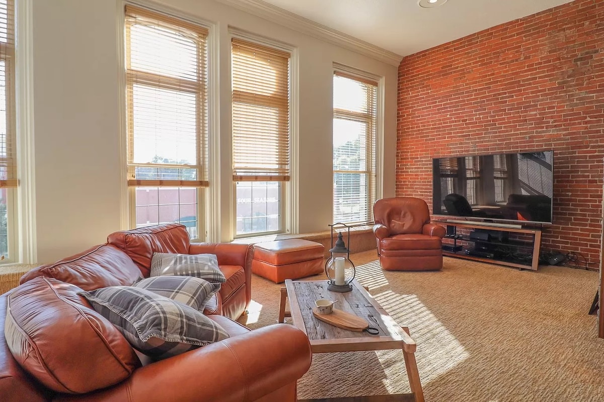 A comfortable living space features a sectional leather sofa, a wooden coffee table, and a cozy armchair. Natural light floods through large windows with blinds, highlighting the warm tones of the brick wall. A flat-screen TV is positioned on a console against the opposite wall.