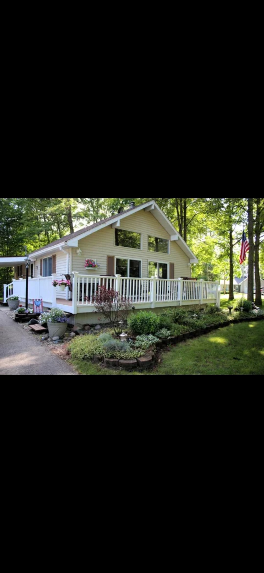A spacious chalet-style cottage is framed by lush greenery and colorful floral arrangements. The porch features railing adorned with hanging plants, while large windows allow natural light to fill the interior. A pathway leads up to the entrance, accompanied by a flag gently waving in the breeze.