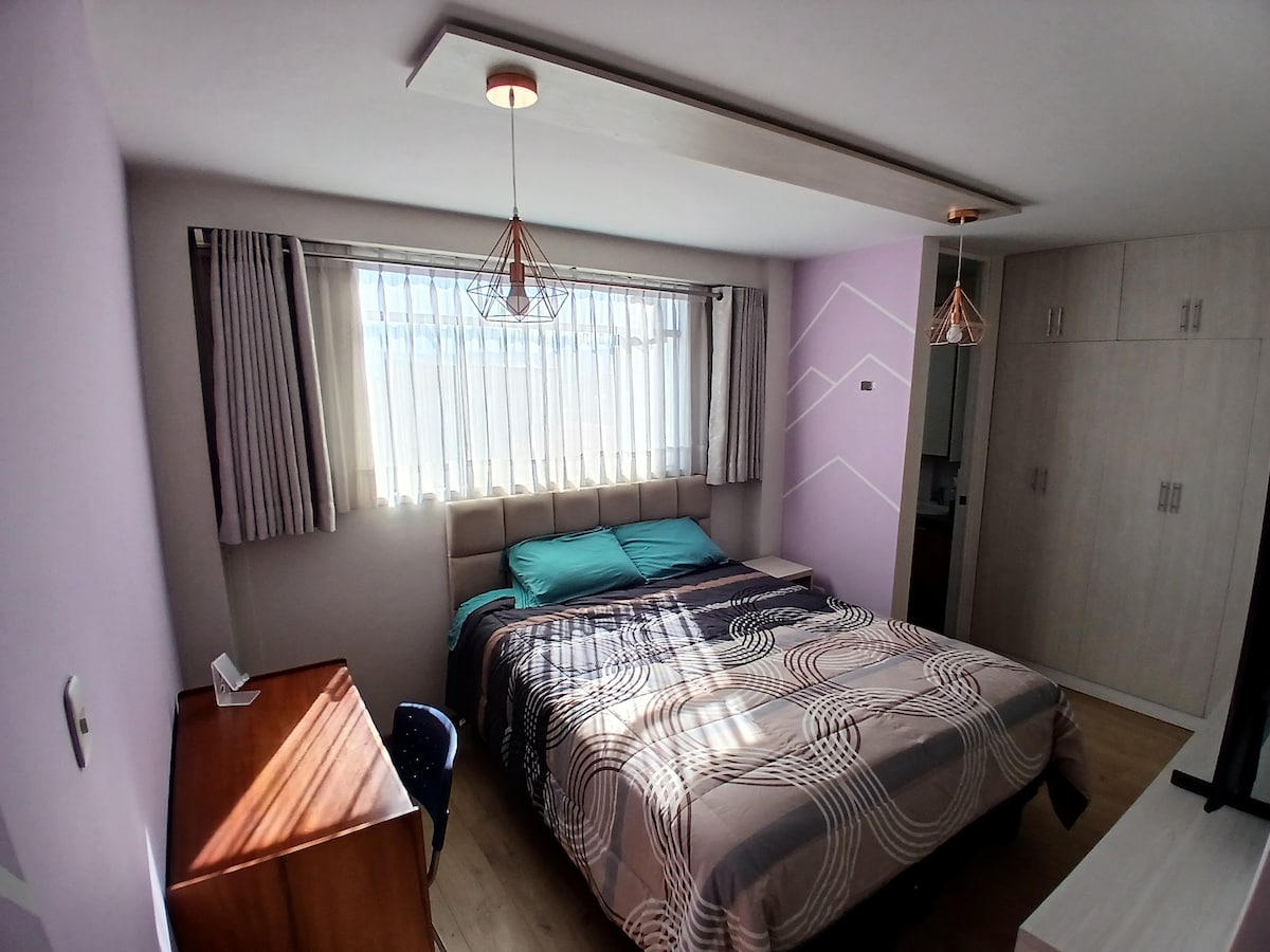 A well-lit bedroom features a spacious bed with a patterned blanket. Two turquoise pillows rest on top. A wooden desk with a chair is positioned beside the bed. Floor-to-ceiling curtains diffuse natural light from the window. Pendant lights add a modern touch to the space.