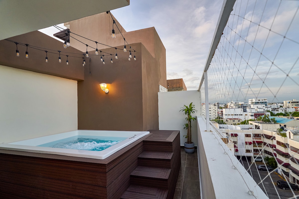 A rooftop terrace is portrayed, featuring a Jacuzzi surrounded by wooden decking and low steps. Warm light is provided by a wall-mounted fixture, and string lights are hung overhead. The view reveals a spacious urban landscape with buildings and greenery in the distance.