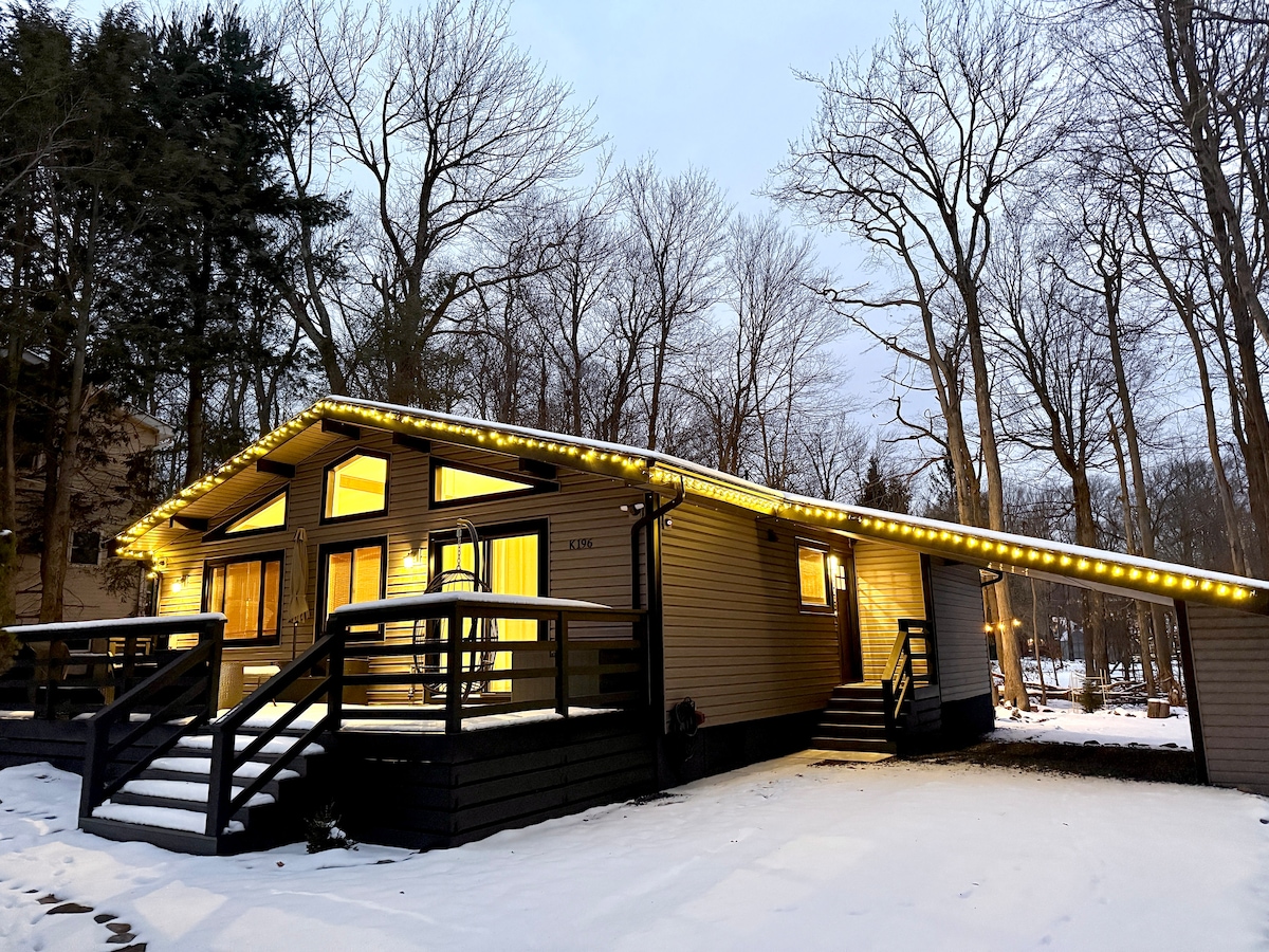 The exterior of the house showcases a modern design with warm light spilling from large windows. Accent lighting along the roof illuminates the home, set against a backdrop of bare trees and a gentle layer of snow covering the ground.