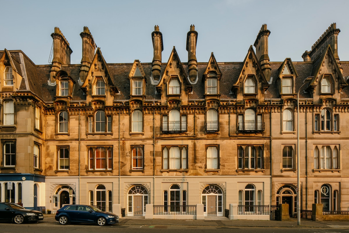 A historic building with a distinctive architectural design is presented, characterized by ornate stonework and multiple gabled roofs. The facade features a mix of arched and rectangular windows, offering a glimpse of character from the early 20th century. A street view shows parked vehicles in front.