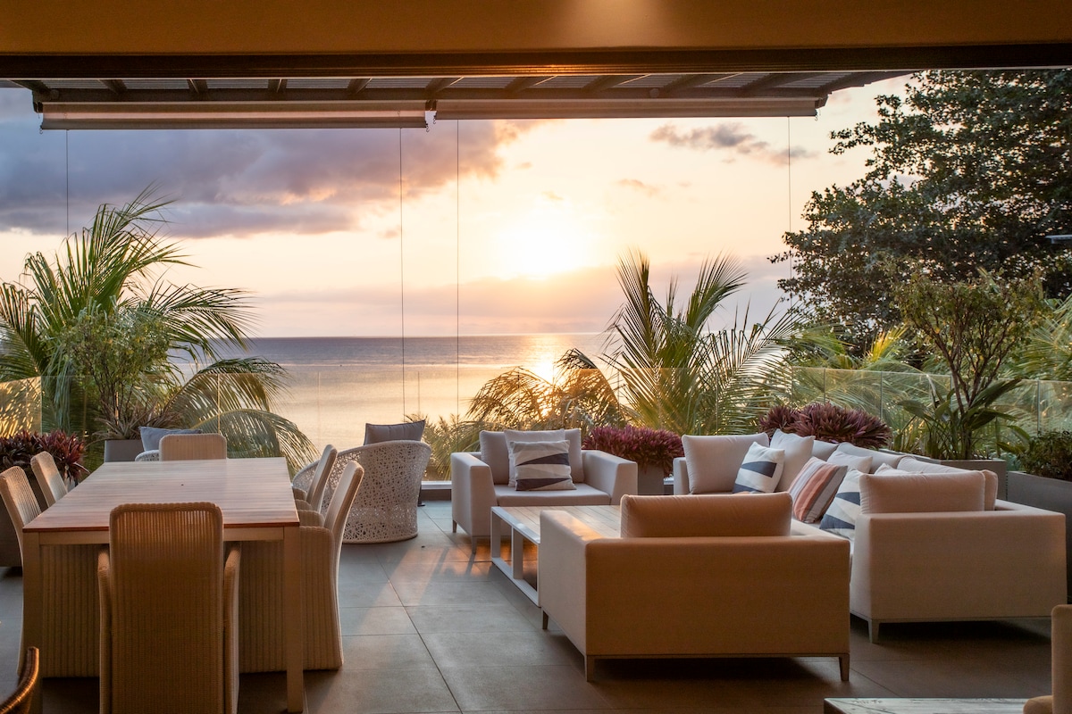 A spacious outdoor terrace features modern, comfortable seating arrangements and a large dining table. Lush greenery surrounds the area, and the ocean can be seen in the background, with a soft sunset casting warm hues across the sky.