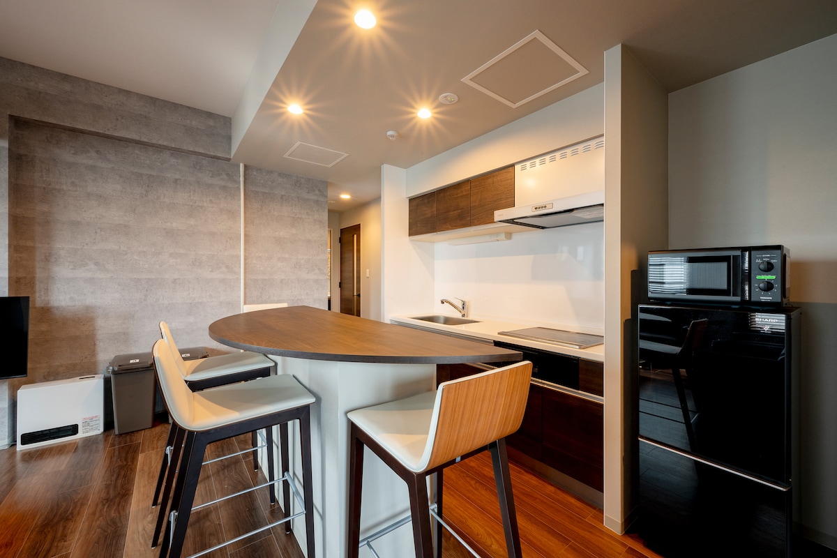 A modern kitchen area is visible, featuring wood cabinets and sleek appliances. A central island with high-backed chairs offers a casual dining space. Soft lighting enhances the warm tones of the wood flooring and cabinetry. The kitchen includes a stove, oven, and refrigerator.