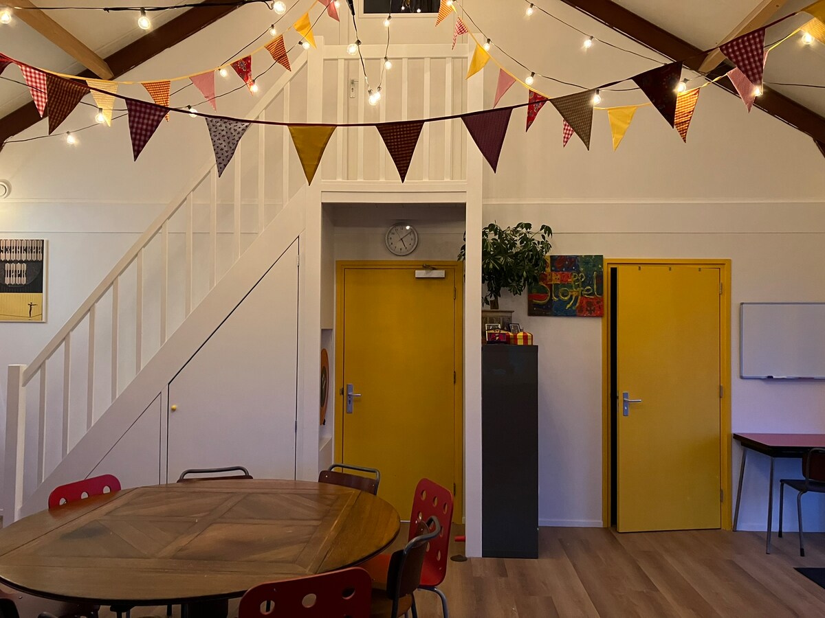 A communal area displays a warm and inviting ambiance, featuring yellow doors and a decorative banner strung overhead. A large, round wooden table is surrounded by red chairs. A staircase in the background leads to an upper level, while a clock and potted plant add functional touches.