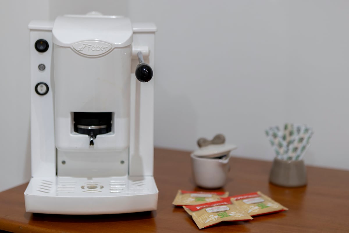 An espresso machine in white is positioned on a wooden table, accompanied by several packets of coffee. A small milk jug and a container with spoons are nearby, creating a well-equipped coffee corner for guests.