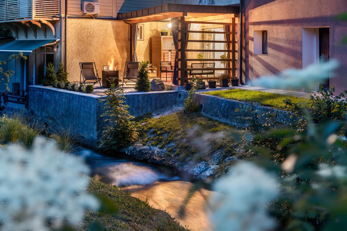 A serene outdoor seating area features two lounge chairs arranged near a fire pit, surrounded by landscaped greenery and soft lighting. The gentle flow of the Jezernica creek can be seen nearby, enhancing the tranquil setting of the terrace.