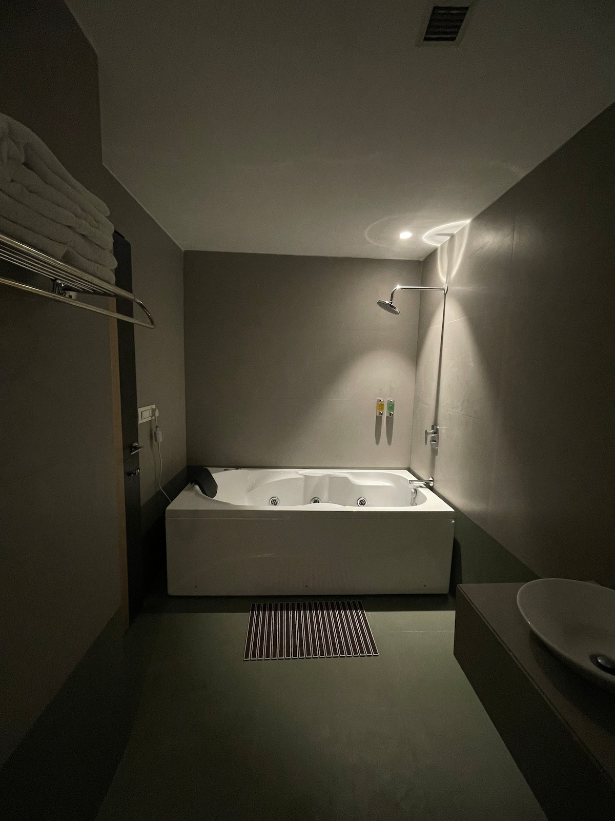 A spacious bathroom features a modern Jacuzzi tub, designed for relaxation. Soft white towels are neatly arranged on a shelf nearby. A sleek showerhead is mounted on the wall, and a minimalist sink is positioned beside the tub, contributing to the room's contemporary design.