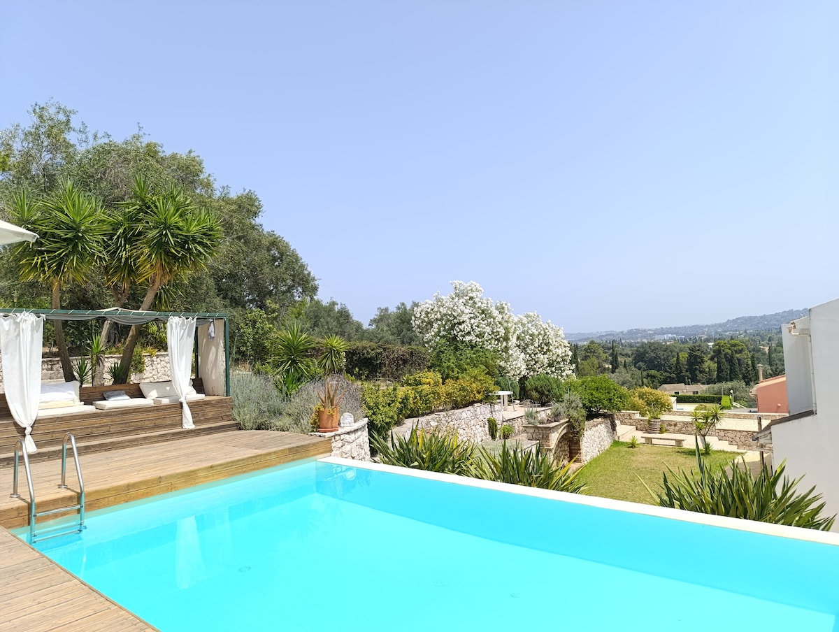 A tranquil outdoor area features a swimming pool with a wooden deck. Lush greenery surrounds the pool, complemented by palm trees and flowering plants. A shaded lounging area with draped fabric provides a spot for relaxation, enhancing the serene view of the landscape.