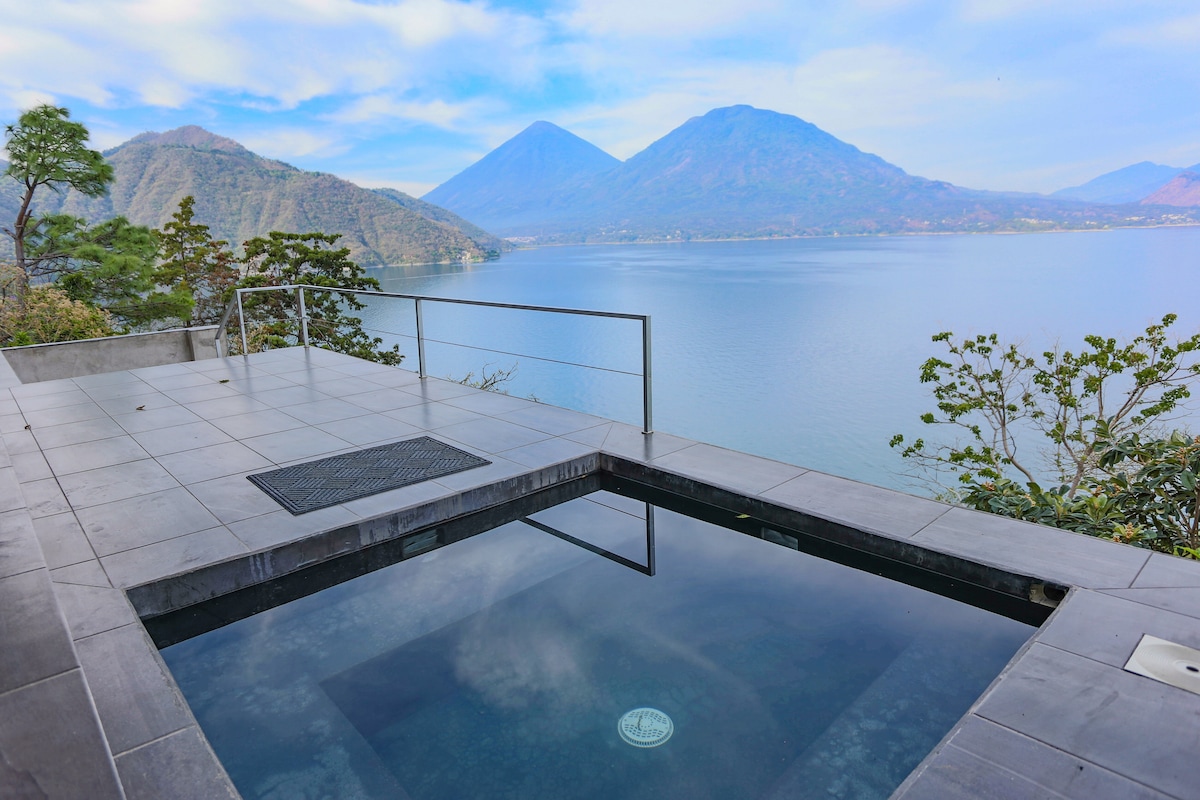 A private outdoor jacuzzi is positioned overlooking the expansive view of Lake Atitlán, framed by lush hills and mountains. The tranquil water reflects the surrounding landscape under a soft, cloudy sky, providing a peaceful atmosphere for relaxation.