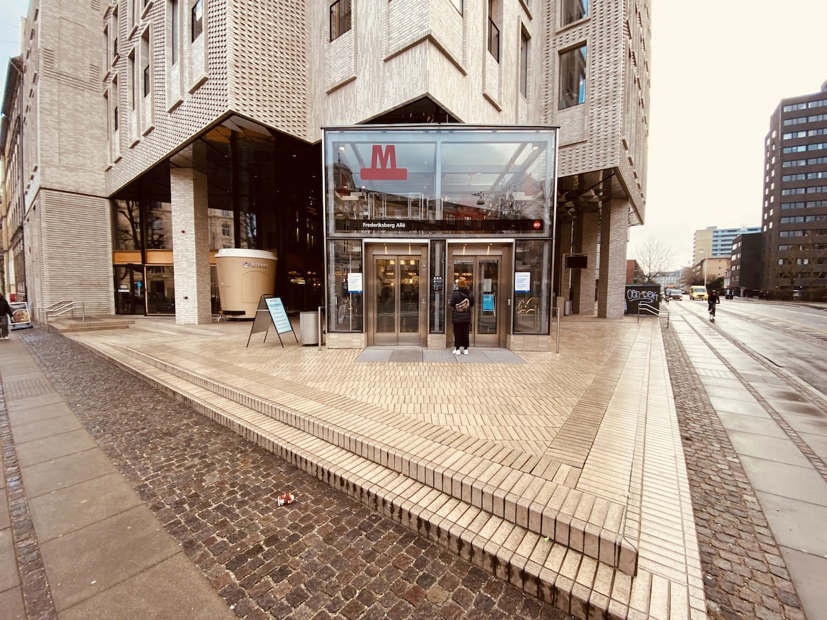 The exterior of a modern building is presented, featuring two large glass entrance doors marked with an 'M' symbol. A person is positioned in front of the entrance, and the surrounding area is paved with light-colored bricks, providing a spacious feel.