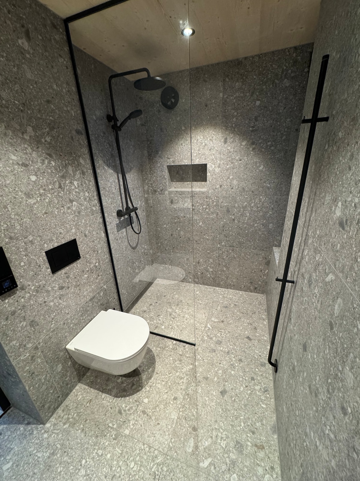 A modern bathroom features polished stone walls and flooring in various shades of gray. A frameless glass shower enclosure is seen, showcasing a sleek rain showerhead and minimalistic controls. A wall-mounted toilet is positioned nearby, contributing to the spacious, clean design.