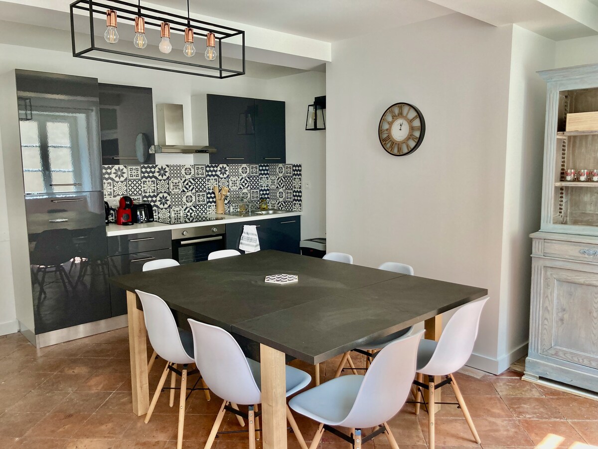 A modern dining area features a large rectangular table surrounded by six white chairs. The kitchen is visible in the background, showcasing sleek black cabinetry and patterned backsplash. A vintage clock adorns the wall, enhancing the room's design, along with a pendant light overhead.