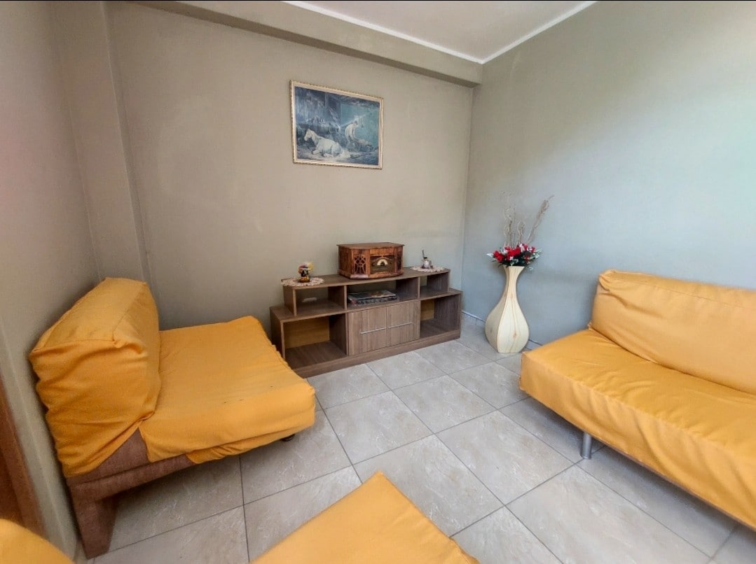 A modest living area is presented with two yellow upholstered sofas and a matching armchair. A wooden media unit is positioned against the wall, featuring decorative items and a floral arrangement above it. A piece of wall art adds a touch of character to the room.